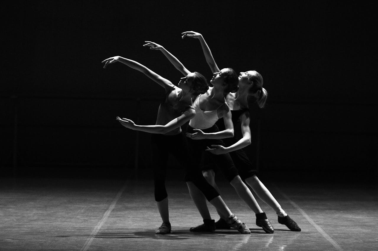 about-us Three female ballerinas perform a synchronized dance on stage in dramatic lighting.